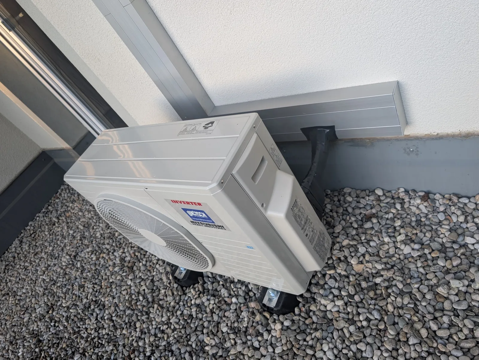 Weißes Klima-Außengerät auf Kies an einer hellen Hauswand