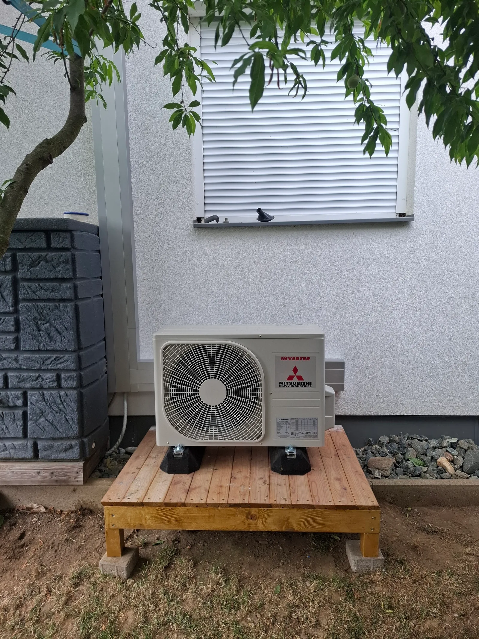 Mitsubishi-Außengerät auf einem Holzpodest im Garten unter einem Fenster